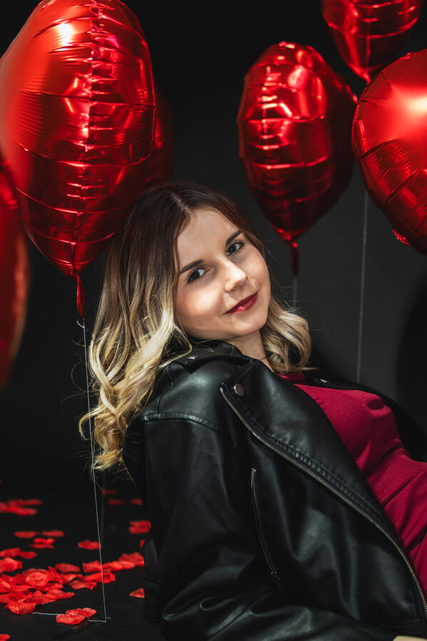 Portret Studio Walentynki Portret kobiety w czerwonej sukience i czarnej kurtce wśród czerwonych balonów w kształcie serc.